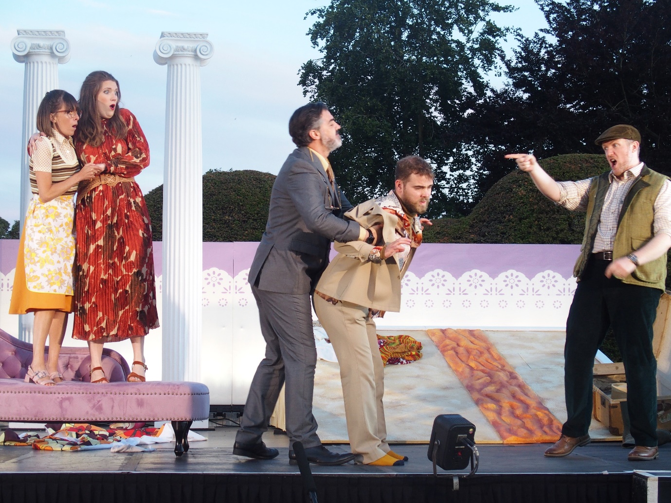 A scene from the opera. One man restrains another at the centre of the stage. On the right hand side of the image, another man gestures accusingly. Two women, standing between columns at the left of the image, look on in horror.